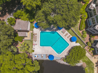 Aerial view of a turquoise community swimming pool with blue umbrellas and lounge chairs, surrounded by leafy oak trees, residential buildings, and a small pond