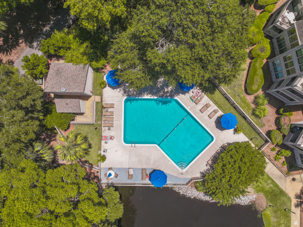 Aerial view of a turquoise community swimming pool with blue umbrellas and lounge chairs, surrounded by leafy oak trees, residential buildings, and a small pond