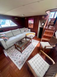 Cozy yacht salon — light gray sectional with colorful pillows, rattan glass coffee table on a seashell rug, wood-paneled walls, nautical clock and a small dog on the stairs.