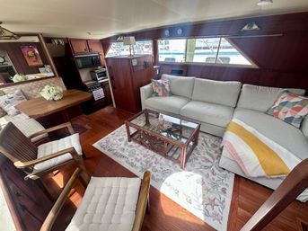 Bright yacht salon interior with light-gray L-shaped sofa, colorful throw pillows and striped blanket, glass-top rattan coffee table on a seashell-pattern rug, wooden dining nook and compact galley with stove and microwave, wood-paneled walls and marina view through long windows.