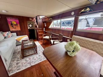 Cozy wood-paneled yacht salon interior at a marina featuring a light-gray sofa, seashell-pattern rug, wicker coffee table, wooden dining table with white hydrangea centerpiece, two wooden armchairs and stairs leading to the deck.
