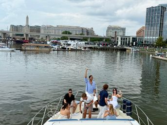 Group of friends enjoying the bow of a yacht at the Washington, D.C. waterfront marina, with waterfront buildings and the Washington Monument visible under a cloudy sky.
