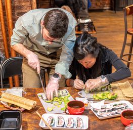 Two people wearing gloves assembling and slicing avocado sushi rolls on a wooden table with bamboo mats, avocado slices, sushi trays and mugs in a hands-on sushi-making workshop.