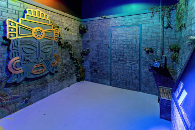 Themed indoor adventure room with textured stone walls, large stylized Mesoamerican mask wall art, creeping vines, camouflaged door and props under moody blue-green lighting.
