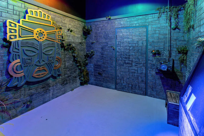 Themed indoor adventure room with textured stone walls, large stylized Mesoamerican mask wall art, creeping vines, camouflaged door and props under moody blue-green lighting.