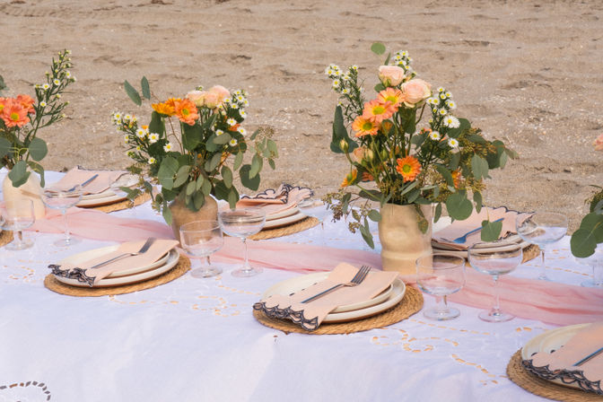 Beach picnic table on sand with white tablecloth and blush runner, wicker placemats, stacked plates, scalloped napkins, coupe glasses, and peach‑orange floral centerpieces in rustic vases