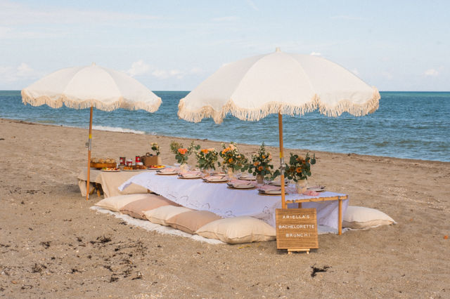 Boho oceanfront beach brunch: low white-draped table with beige cushions, fringed umbrellas, floral centerpieces and place settings set on a sandy shore by turquoise water.