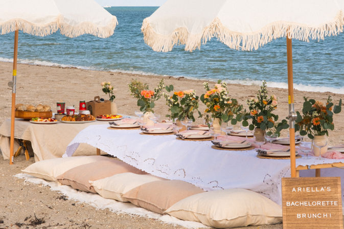 Seaside brunch setup on a sandy beach: low linen-draped table with pastel place settings and wicker chargers, floral centerpieces, fringed umbrellas, floor cushions and a letter board announcing a bachelorette brunch.