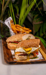 Sesame-seed sandwich with a runny fried egg, melted cheddar, tomato and greens on a wooden board, served with a metal basket of golden crinkle-cut fries — brunch cafe style.