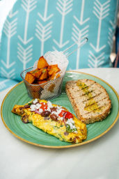 Brunch plate with Mediterranean-style omelette topped with feta, olives and cherry tomatoes, grilled multigrain toast and seasoned potato cubes in a metal basket on a bright café table.