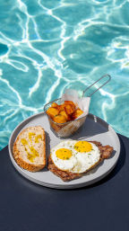 Poolside breakfast plate with steak topped by two sunny-side-up eggs, multigrain toast drizzled with olive oil, and crispy seasoned potato cubes in a mini fryer basket beside a bright blue swimming pool