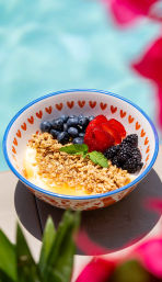 Poolside yogurt bowl with granola and honey, topped with blueberries, sliced strawberries, blackberries and a mint leaf in a heart‑trimmed bowl