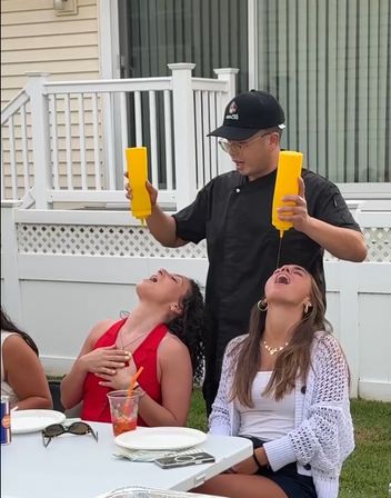 Outdoor backyard party: man in a black shirt pours from two yellow squeeze bottles into the open mouths of two seated women at a picnic table in front of a white fence, with plates and drinks on the table.
