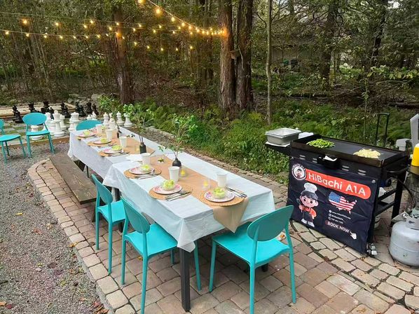 Outdoor garden dinner setup on a stone patio: long table with white tablecloth, turquoise chairs, woven place settings and salads, string lights overhead, portable griddle cooking vegetables beside a propane tank, wooded backdrop and oversized outdoor chess pieces.