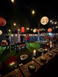 Festive backyard nighttime dinner with a long table set for guests, candles and place settings, an outdoor hibachi grilling station, and glowing string lights with red-and-white paper lanterns over the lawn.