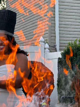 Vibrant orange flames from a backyard fire pit in front of a smiling person wearing a tall black hat and white strapless top, with a suburban house, white fence and shrubs in the background.
