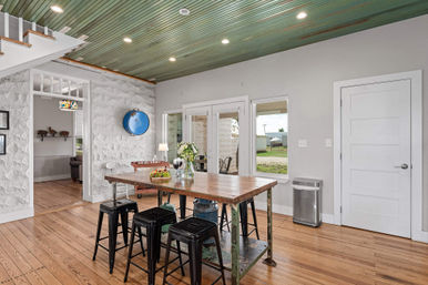 Sunlit farmhouse-style dining area with a reclaimed wood table and black metal stools, green tongue-and-groove ceiling, white textured stone accent wall, hardwood floors, glass patio doors and window, and a vase of fresh flowers as a centerpiece.
