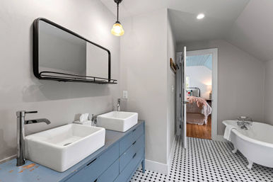Bright modern farmhouse bathroom with blue double vanity and rectangular vessel sinks, black-framed mirror, black-and-white hexagon tile floor, freestanding white clawfoot tub, and a bedroom visible through an open door.