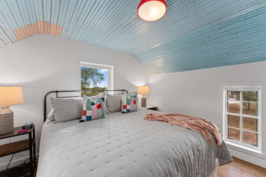 Cozy attic bedroom with sky-blue beadboard ceiling, large white quilted bed with colorful geometric pillows, bedside lamps, wood floor and small windows with tree views