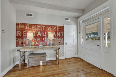 Inviting farmhouse entryway with white front door and sidelights, rustic wood console table with two glass lamps, large distressed red vintage sign, wicker storage basket and warm hardwood floors.