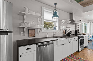 Cheerful modern farmhouse kitchen with white cabinets, black countertops, stainless steel appliances, apron-front farmhouse sink under a window, open white shelving with glassware, blue industrial pendant light, hardwood floors and colorful woven rug.