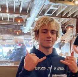 Young blond man in a dark hoodie flashing double shaka hand signs inside a rustic industrial café with exposed ductwork, wooden beams and hanging pendant lights.