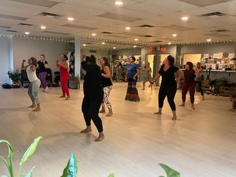 Energetic group dance class of women dancing barefoot in a bright mirrored studio with light wood floors, string lights and plants — community fitness/dance session.