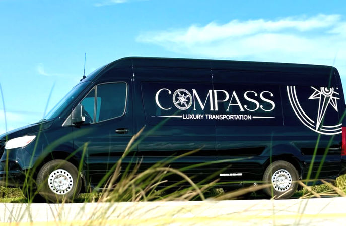 Sleek black luxury shuttle van with a large compass-style logo parked on a sunny coastal road, dune grasses in the foreground and blue sky overhead.