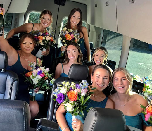 Seven smiling bridesmaids in teal dresses holding colorful bouquets and a raised champagne bottle while celebrating inside a wedding shuttle van.
