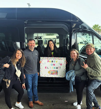 Five smiling adults posing by a black shuttle van at a curb, woman in the center holding a colorful sign that reads “PARTY ON ELAINE!!” — friends ready for a fun group celebration and ride.