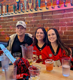 Three cheerful friends at a sports bar with exposed brick and a row of beer taps overhead; two wearing red Georgia Bulldogs jerseys, pitcher, cocktails and beer cups on the table.