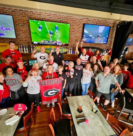 Cheering group of Georgia Bulldogs fans in red and black holding a large Georgia Bulldogs banner inside a brick-walled sports bar, watching college football on multiple TVs.