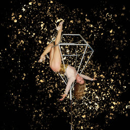 Glam aerial performer in a sequined bodysuit and heels hangs upside down from a metallic geometric aerial cube on a pole against a black backdrop splashed with gold confetti.