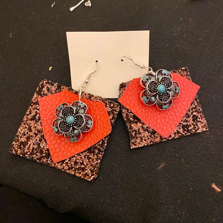 Handmade boho dangle earrings with silver flower charms and turquoise centers layered on red faux-leather shapes and rose-gold glitter square backings, photographed on black fabric.