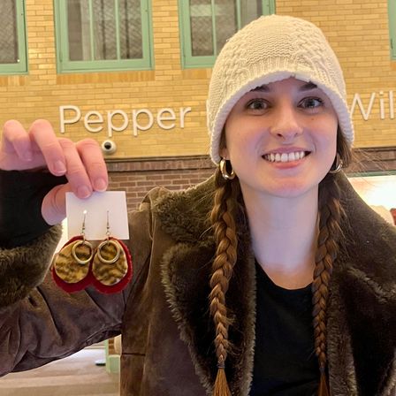 Smiling person in a knit beanie and braided hair holding handmade red-and-leopard-print dangle earrings inside a brick-walled indoor craft market