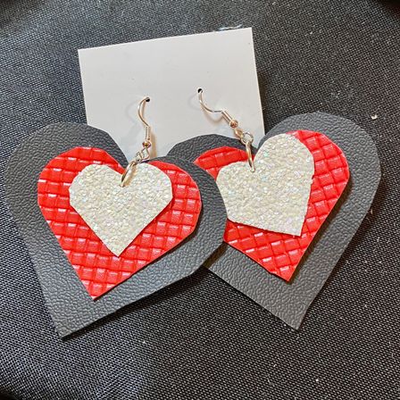 Handmade layered heart earrings — black faux-leather base, textured red middle heart, sparkly white glitter top heart with silver hooks on a white display card, styled on black fabric; artisan Valentine’s Day statement earrings, gift-ready