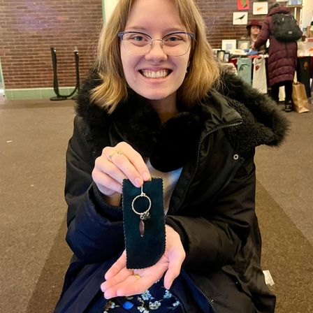 Smiling person at an indoor craft fair holding a handmade silver hoop pendant with tiny charms and a teardrop stone displayed on a dark velvet strip, wearing a black winter coat.
