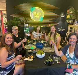 Five smiling women raising drinks around a table at an indoor plant workshop, assembling succulents and terrariums with bowls, moss and tools; neon circular wall sign, wood-slat accent wall and potted plants in the plant studio background.