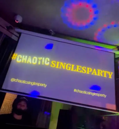 Projection screen displaying bold yellow hashtag '#CHAOTICSINGLESPARTY' at a lively singles party, colorful disco light rings on the ceiling and a silhouetted person in the foreground.