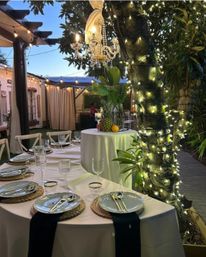 Evening garden patio dining with white-linen round tables, woven placemats, gold flatware and wine glasses, twinkling string lights wrapped around a tree, hanging crystal chandelier and tropical pineapple centerpiece — romantic outdoor event vibe.