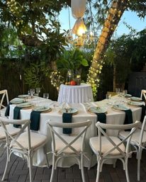 Cozy outdoor garden dinner setup with white-linen round table, green napkins, rattan chargers, pale blue plates and glassware, white cross-back chairs under a chandelier and tree-wrapped string lights.