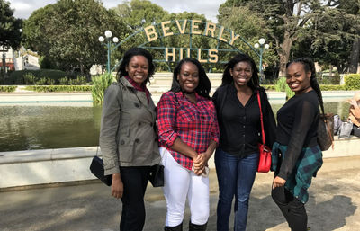 Four smiling friends posing by the iconic Beverly Hills sign and reflecting pool with park trees behind them