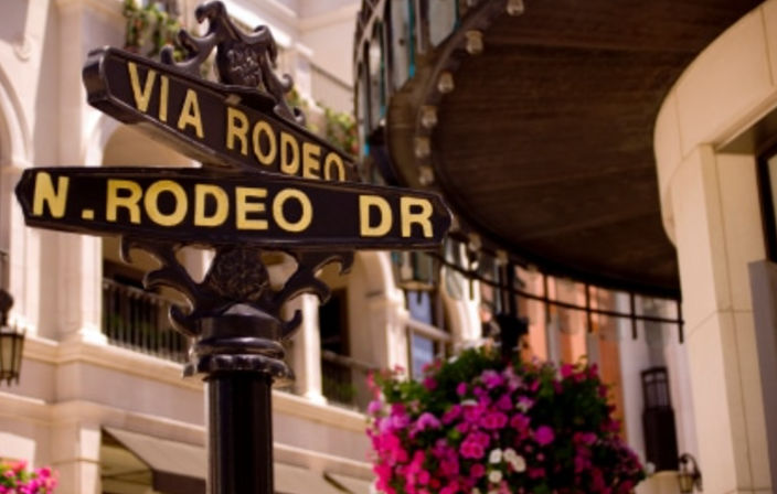 Black-and-gold street signs reading "Via Rodeo" and "N. Rodeo Dr" on an ornamental post in a chic upscale shopping district, with pink hanging flowers and elegant building facades