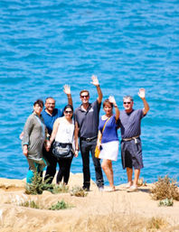 Cheerful group of six people in casual summer clothes standing on a sunny coastal bluff, waving at the camera with bright blue ocean water behind them.