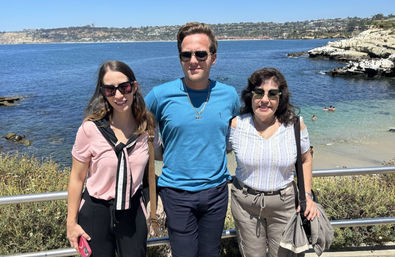 Three adults in sunglasses posing at a sunny rocky coastal overlook with blue ocean, cliffs and a small sandy cove with swimmers visible in the background