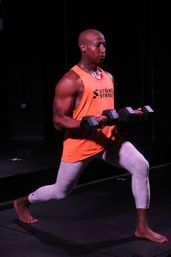 Person performing a weighted forward lunge barefoot on a dark indoor gym studio floor, holding hex dumbbells and wearing a bright orange tank top and light compression leggings — strong, focused fitness pose.
