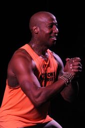Smiling fit person in a bright orange tank top performing a squat with hands clasped, wearing stacked beaded bracelets and a neck tattoo, energetic studio workout against a black background.