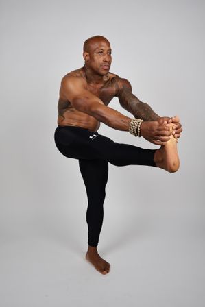 Shirtless tattooed person in black leggings performing a one-legged yoga balance stretch, gripping an extended foot and wearing beaded bracelets against a neutral studio background.