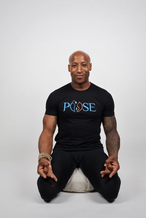 Smiling bald person kneeling on a meditation cushion in a neutral studio, wearing a black T-shirt with a colorful logo, hands in meditation mudra, beaded bracelet and tattooed arms.