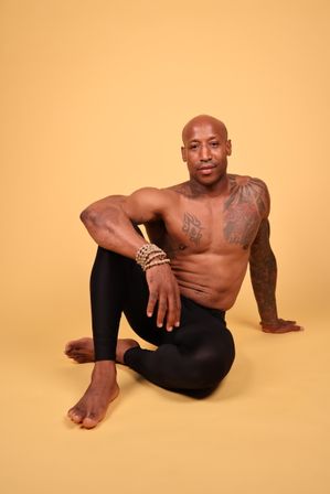 Fit shirtless man with tattoos and wooden bead bracelets seated in a yoga twist pose on a warm yellow studio background, wearing black leggings — yoga and fitness portrait.